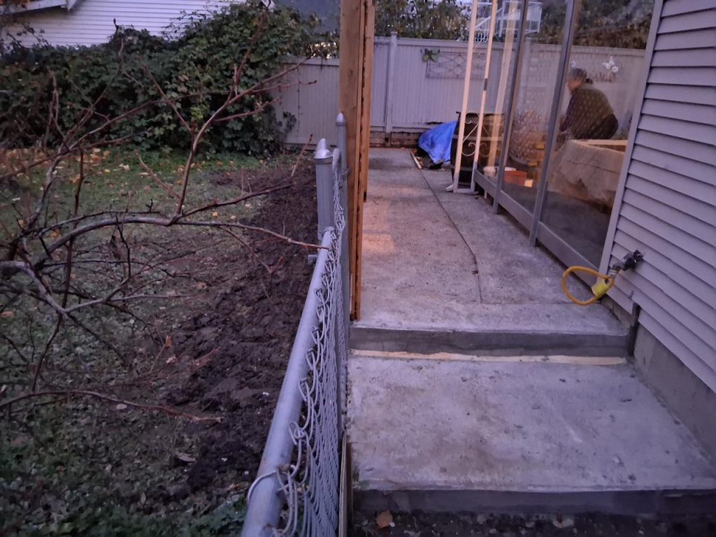 Concrete back patio path beside a house with a sliding glass door; a person sits inside on a couch viewed through the door, and a yellow hose is attached to the siding outside.