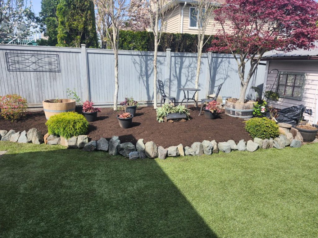 Backyard garden with a mulched planting bed, potted plants, and a small metal table with two chairs in front of a light gray wooden fence; a rock border and lush green lawn in the foreground.