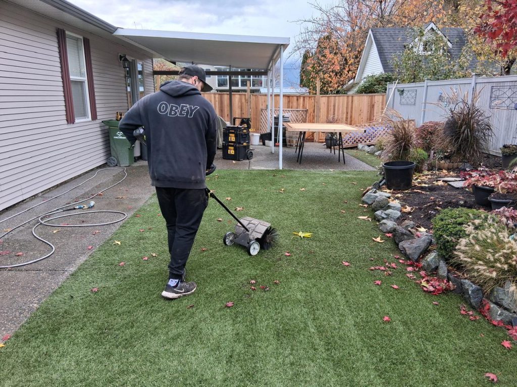 Man in a dark hoodie pushing a wheeled lawn aerator across a backyard lawn district of a house.