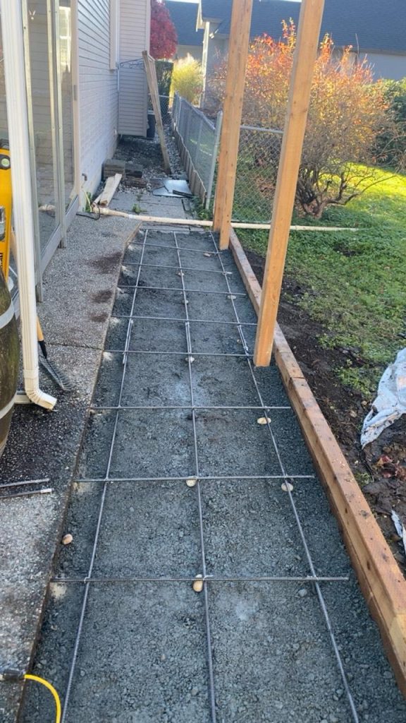 Narrow outdoor walkway under construction with a rebar grid set in packed gravel beside a house, bordered by wooden posts for framing a future fence or deck paneling.