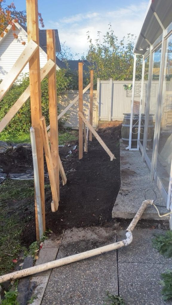 Unfinished wooden fence supports along a backyard path beside a glass-walled house; a dirt trench runs between the fence and the house edge.