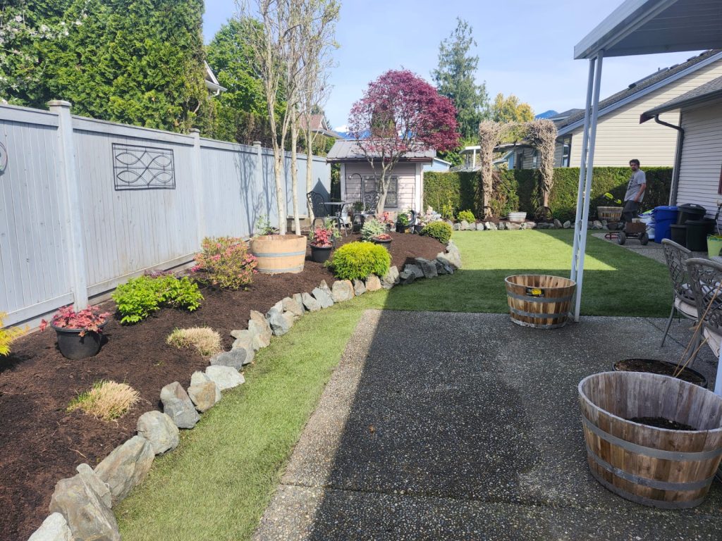 Backyard garden with a rock-edged flower bed along a pale wooden fence and a person standing near the house in the distance.
