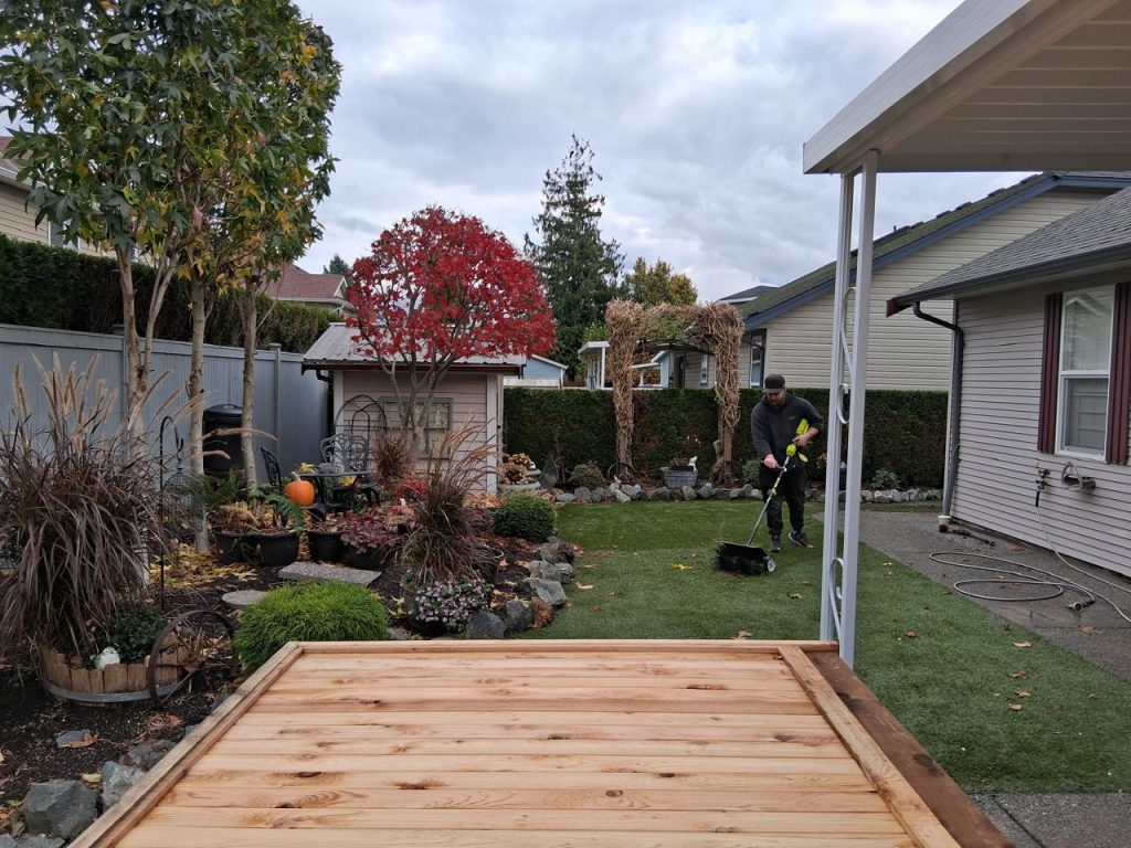 Man pushing a small lawn mower across a suburban backyard with a wooden deck in the foreground and autumn trees in view.