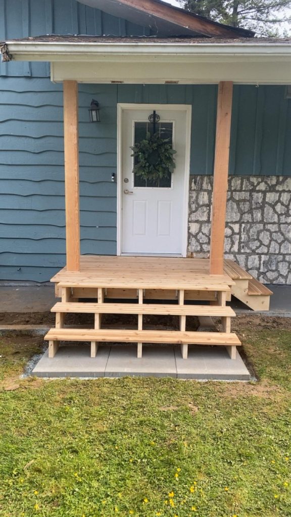 Front porch with newly built wooden steps and posts, blue siding, and a white door with a wreath.