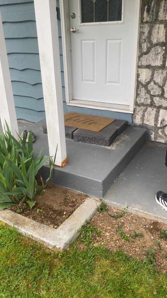 Front door entry with a brown welcome mat on dark gray steps, blue siding and stone wall on the side, and a shoe nearby on the left.