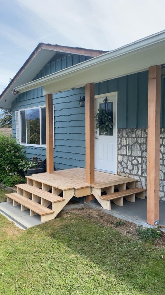 Wooden front porch under construction with three vertical posts, steps, and a wreath-adorned white door on a blue house exterior.
