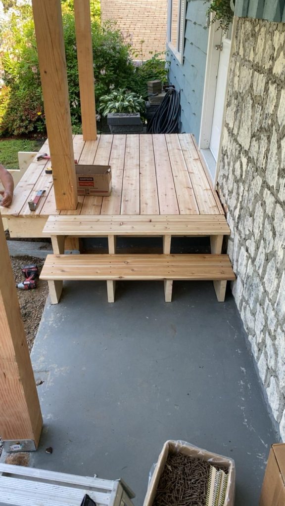 New wooden deck under construction on a concrete patio, with steps and posts ready for railing, tools and a box nearby.