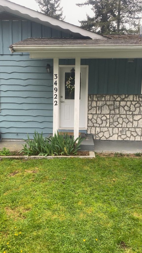 Front entrance of a blue house with a white door and a vertical address plaque showing 34922; wreath on the door, a stone half-wall to the right, and a small garden bed with green plants in front.