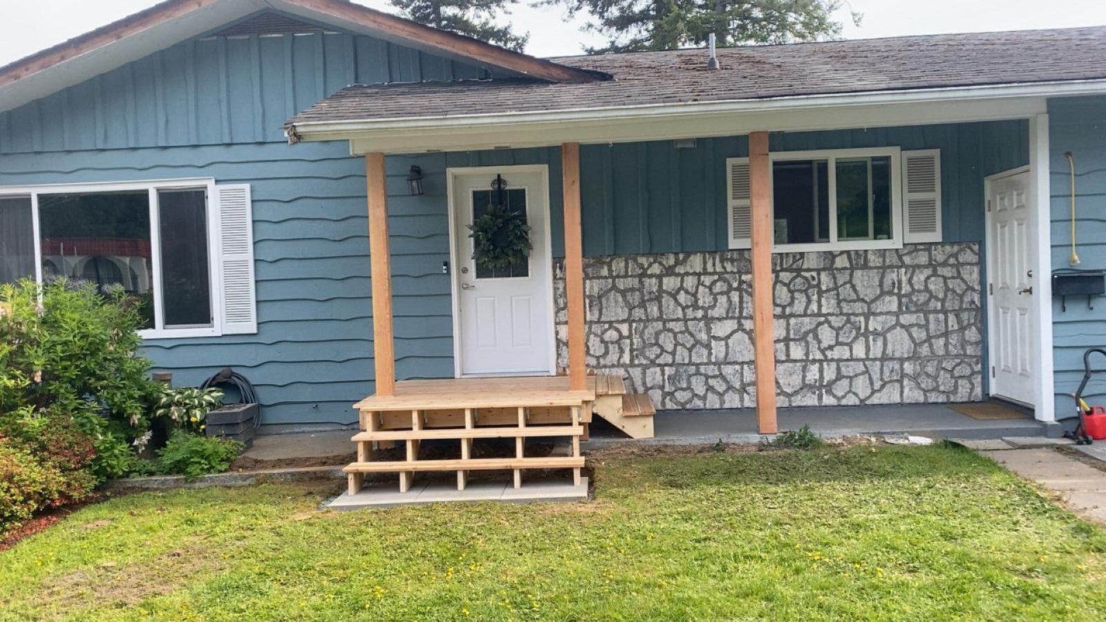 Blue ranch-style house with a small wooden front porch and steps, wreath on the door, and a stone-patterned half-wall under the window.