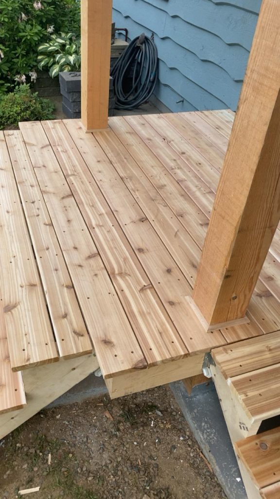 Unfinished wooden deck with vertical posts over concrete footings, surrounding a garden bed and blue siding; a coiled hose sits in the corner behind the posts.