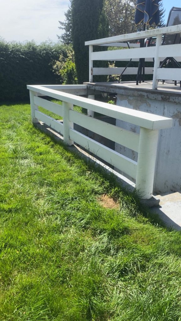 White wooden railing surrounds a small raised patio with outdoor furniture in a grassy backyard setting.