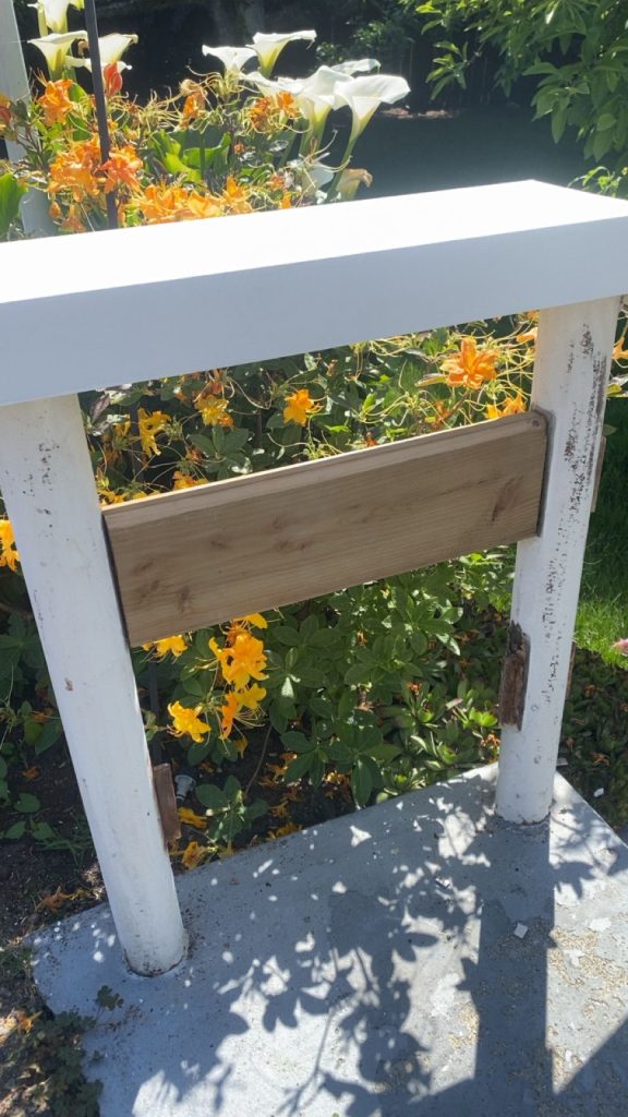 White-painted railing and weathered wood panel in a garden, with orange-yellow flowers and green leaves behind it in bright sunlight.
