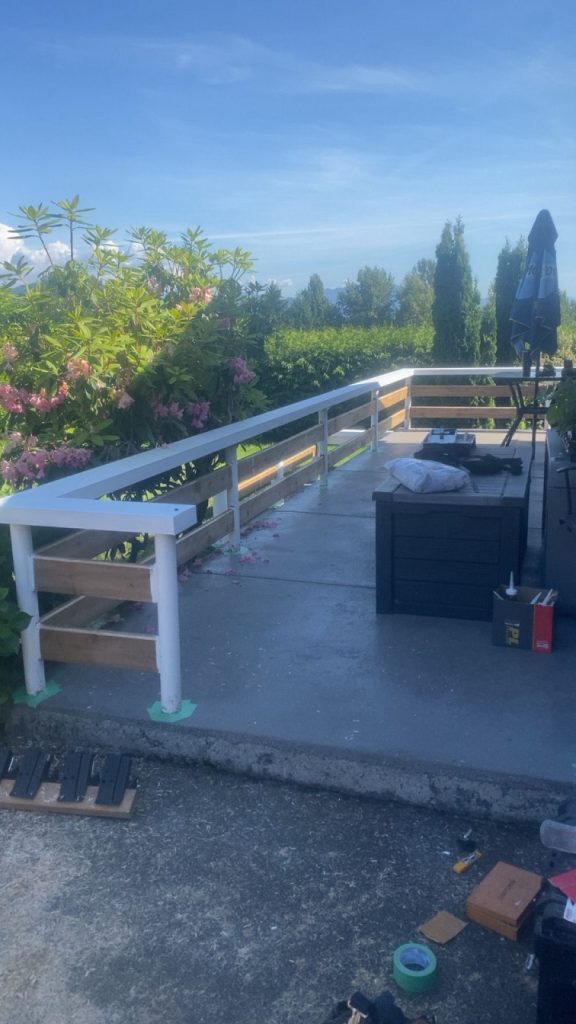 Outdoor patio or deck with white railing and wooden steps, under a blue sky with green plants and pink flowers nearby, tools scattered on the floor.