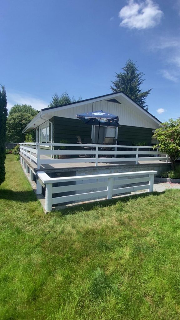 Green single-story house with white rail-deck and outdoor seating under a blue umbrella on a sunny day