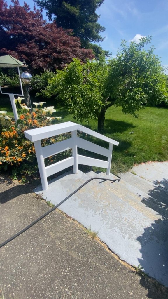 White wooden railing around a small outdoor stairway in a sunny garden with orange flowers and trees nearby.