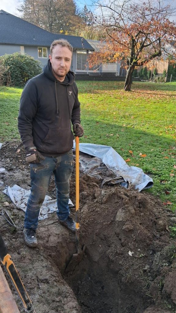 Man in a black hoodie and gloves stands over a dug hole in a backyard, gripping a shovel; dirt heap and tarp nearby, autumn leaves on the grass.