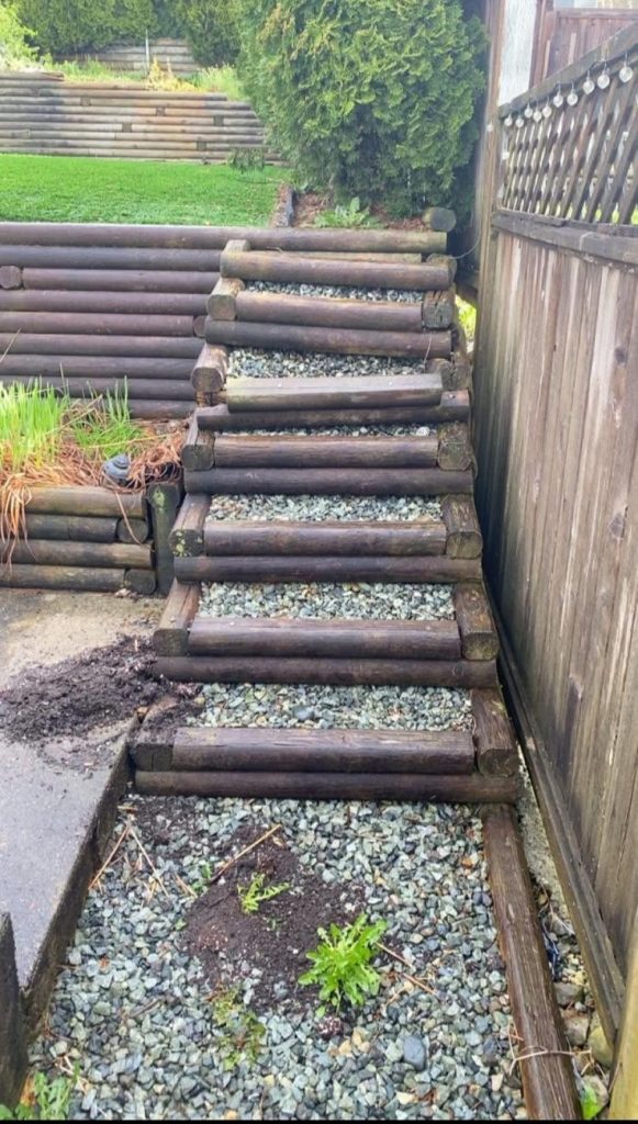 Wooden log-stair staircase built into a gravel path between a fence and a garden, with stacked timber in background.