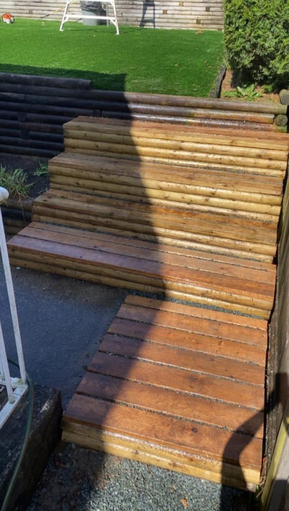 Stack of wooden steps outdoors beside a wooden fence and green lawn, with a large shadow crossing the steps.