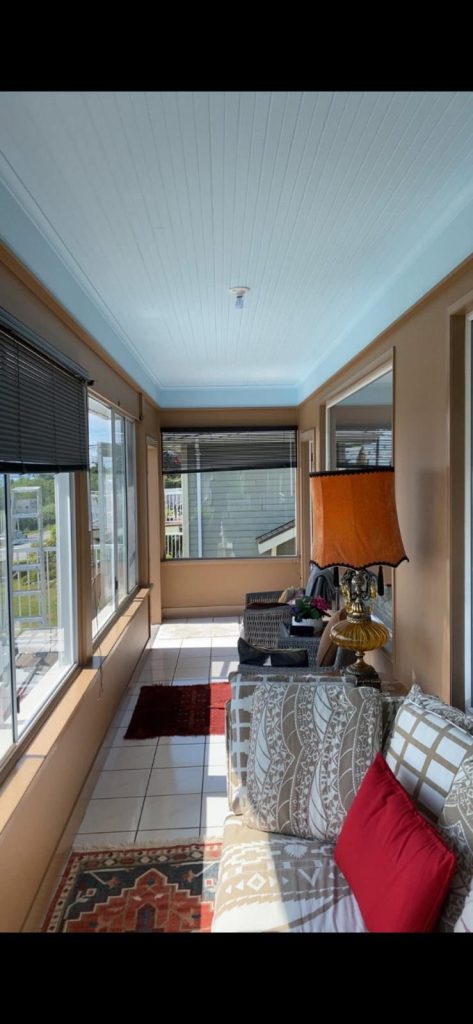 Sunlit enclosed porch with large windows and blinds, beige walls, and a patterned sofa with a red throw pillow, beside an ornate orange lampshade.