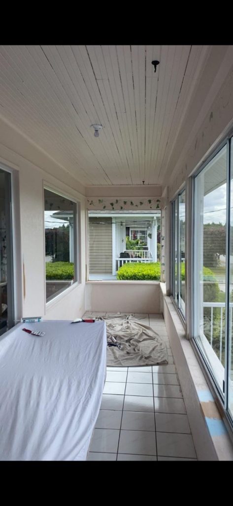 Bright enclosed porch with large windows, a table covered by a white cloth, and a beige drop cloth on the tiled floor with tools on the table reliefs outside visible through the windows.