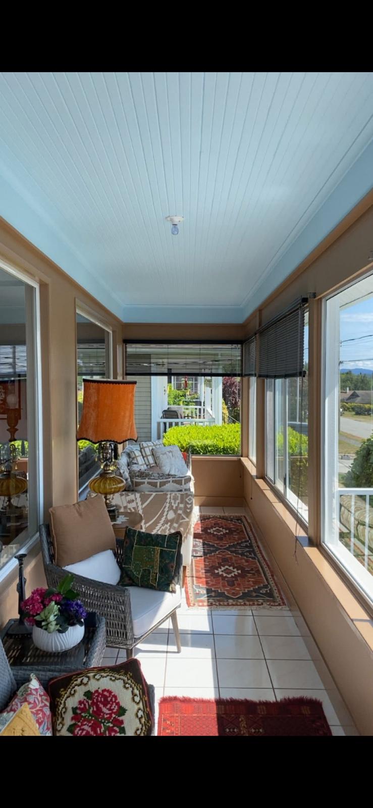 Bright enclosed porch with wicker seating, colorful cushions, and a patterned rug along glass windows overlooking a garden.